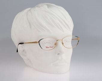 May include: Gold-tone metal spectacles with oval-shaped rims featuring a dark patterned accent on the outer edges. The right lens displays the red text Paloma Picasso. The thin temples transition to black tips, resting on a white mannequin head.