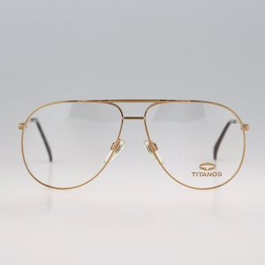 May include: Gold-toned aviator-style eyeglasses with a thin metal frame and a double bridge. The right lens features the brand name TITANOS printed in gold lettering. The glasses have clear lenses, adjustable nose pads, and black temple tips.