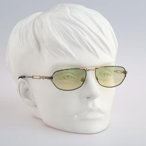 Puede incluir: Gafas de sol con montura dorada y lentes tintados de verde. Las gafas tienen un diseño detallado en la montura y una patilla negra.