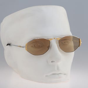 Puede incluir: Un par de gafas de sol con montura dorada y lentes marrones sobre una cabeza de maniquí blanca.