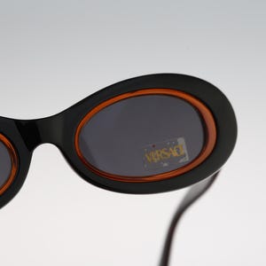 May include: Black oval sunglasses with orange frames and dark lenses. The sunglasses have the word "VERSACE" printed on the lenses.