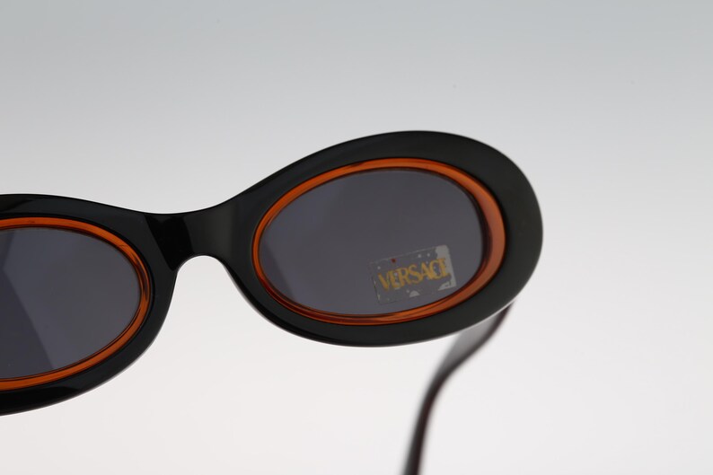 May include: Black oval sunglasses with orange frames and dark lenses. The sunglasses have the word "VERSACE" printed on the lenses.