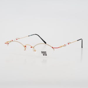 May include: Gold-toned rimless eyeglasses with oval lenses and red and gold decorative accents. The temples are gold with black ear pieces. The text "DOLCE VITA" is printed on the lenses.