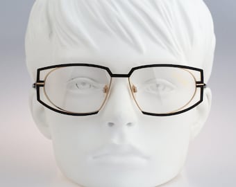 May include: Eyeglasses featuring a dual-layered frame design with a matte black outer geometric structure and a thin gold-toned inner rim. The lenses are clear and transparent, held within a structured metal frame that sits on a white mannequin head.