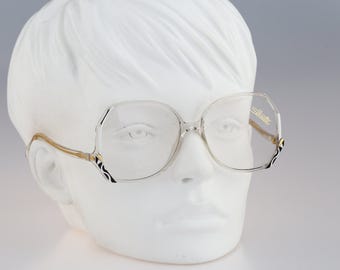 May include: Clear oversized eyeglasses with a gold-tone frame and black and white patterned accents at the hinges. The lenses feature the Silhouette brand logo in gold lettering on the upper right corner, displayed on a white mannequin head.