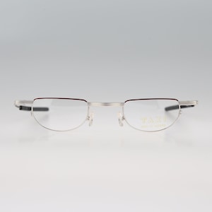 May include: Silver-rimmed glasses with a red accent on the top of the frame. The glasses have a rimless design and say "TAXI Made in Japan" on the right lens.