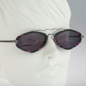 Puede incluir: Un par de gafas de sol plateadas con una montura geométrica facetada. La montura está estampada con rayas rosas y azules. Las lentes son de color gris oscuro.