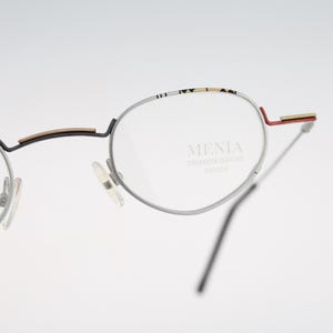 Puede incluir: Gafas de metal plateado con monturas redondas, borde superior estampado, puente negro con detalle dorado y puntas de patilla rojas. El cristal derecho tiene grabado el texto MENIA, seguido de un texto más pequeño y menos legible.