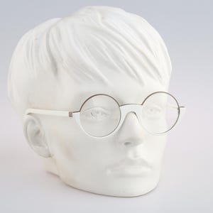 May include: White and brown-rimmed round eyeglasses on a white mannequin head. The glasses have a minimalist design with a thin frame and clear lenses. The mannequin head is a neutral backdrop, highlighting the eyewear.