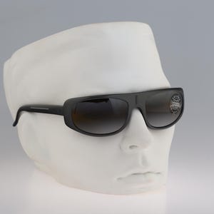 May include: Black sunglasses with a thick frame and gradient lenses featuring a circular sticker on the right lens. The eyewear is displayed on a white mannequin head, showcasing a wrap-around style with a solid bridge and dark temples.