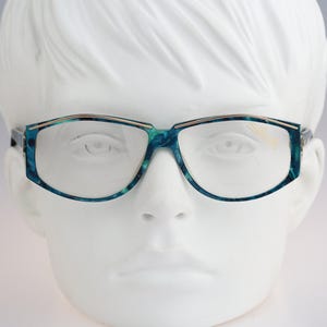 Könnte beinhalten: Brille mit einem blau-türkisfarbenen Marmormuster auf dem Rahmen. Die Brille hat eine geometrische Form mit einem dünnen goldfarbenen Metallakzent entlang der Oberkante der Gläser. Die Gläser sind klar und der Rahmen wird auf einem weißen Kopfmodell gezeigt.