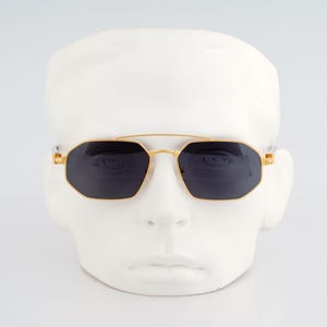 Puede incluir: Gafas de sol con montura dorada y lentes oscuras. Las gafas de sol tienen un doble puente y una forma geométrica. Las gafas se exhiben en una cabeza de maniquí blanca sobre un fondo blanco.
