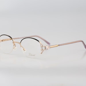 May include: Gold-tone rimless eyeglasses with a black frame and clear lenses. The glasses have a decorative detail on the side with rhinestones and the word "Diva" printed on the right lens.