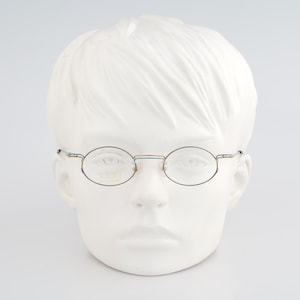 May include: Round eyeglasses with a thin, silver-colored metal frame. The glasses are displayed on a white mannequin head. The lenses are clear. The overall style is minimalist and classic.