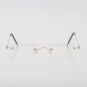 Pode incluir: Óculos sem aro com uma ponte e componentes de armação de metal fino em tom prateado. O design inclui almofadas de nariz ajustáveis e hastes de metal finas que terminam em pontas de plástico preto, proporcionando um visual leve e elegante.