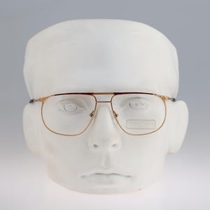May include: Gold-rimmed glasses with clear lenses on a white mannequin head. The glasses have a subtle, textured design on the frame.