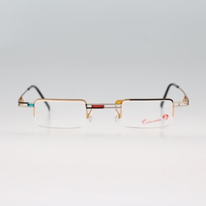 May include: Gold-tone rimless eyeglasses with a black and silver accent on the bridge. The brand name "Casanueva" is printed in red on the right lens.