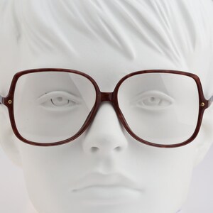 May include: Oversized square eyeglasses with a dark reddish-brown tortoiseshell pattern frame. The glasses feature clear lenses and small gold-toned metal accents at the hinges where the temples connect to the front of the frame.
