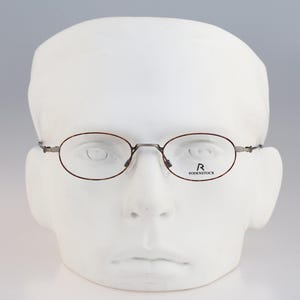 May include: Oval-shaped eyeglasses with a thin, dark brown metal frame. The lenses are clear, and the right lens features the black printed logo and text Rodenstock. The glasses are displayed on a plain white mannequin head, highlighting the frame.
