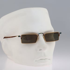 Peut inclure: Lunettes de soleil rectangulaires dotées de fines montures en métal doré et de verres teintés foncés. Les branches et le pont présentent un motif écaille de tortue marron et noir moucheté, contrastant avec les bords dorés de la monture.