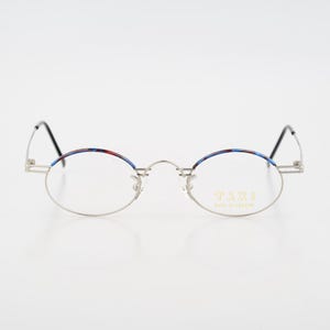 May include: Oval-shaped eyeglasses with a silver frame and black temples. The upper rims of the lenses feature a colorful pattern of red, blue, and white. The text "TAXI MADE IN FRANCE" is visible on the right lens.