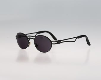 May include: Round sunglasses featuring a matte black metal frame and dark tinted lenses. The eyewear includes thin, angular temples and adjustable nose pads, showcasing a sleek, minimalist construction with a distinct bridge design.