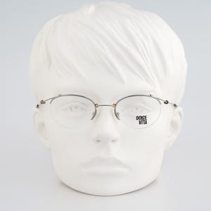 May include: Oval-shaped eyeglasses with a silver frame and clear lenses. The glasses are displayed on a white mannequin head. The text "DOICE VITA" is printed on the right lens. The glasses have small decorative accents on the temples.