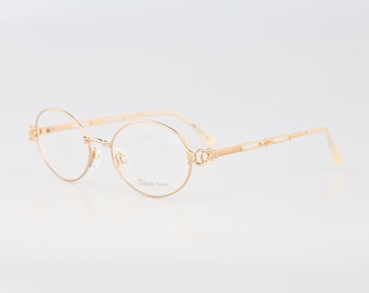May include: Gold-toned metal spectacles with round frames and thin temples. The right lens features the text Diva in a cursive font. The bridge and hinges display intricate decorative detailing, and the temple arms have a repeating open link pattern.