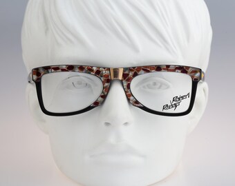 May include: Eyeglasses with a rectangular frame featuring a mosaic pattern of brown, white, and metallic tones on the upper rim and bridge. The lower rims are solid black. The right lens displays the text Robert Rudger in a black script font.