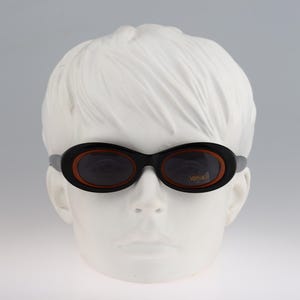 May include: A pair of black oval-shaped sunglasses with brown frames and dark lenses. The sunglasses are on a white mannequin head.
