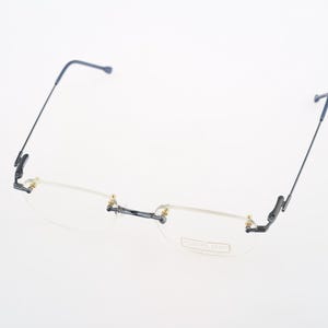 Puede incluir: Gafas sin montura con patillas finas de metal oscuro y un puente con detalles en tono dorado. Las lentes transparentes son rectangulares con bordes redondeados, y la lente derecha muestra un logotipo de marca grabado en la esquina.