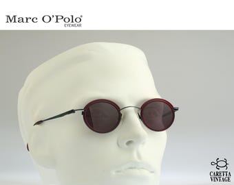 May include: Marc O'Polo eyewear round sunglasses featuring dark red circular rims and thin black metal temples. The lenses are dark tinted, and the frame design includes a bridge connecting the two circular lenses. The text Marc O'Polo Eyewear and Caretta Vintage is visible.