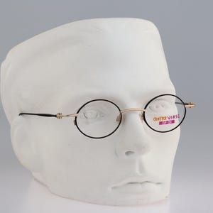 Puede incluir: Gafas redondas con monturas de metal fino en tono dorado y bordes negros. El cristal derecho tiene una pegatina con el texto "CONTRA VERSE" en naranja y morado, con "CLIP-ON" impreso en blanco sobre un fondo morado bajo la marca.