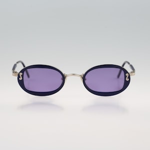 May include: A pair of oval-shaped sunglasses with a silver frame and purple lenses. The sunglasses have a small, decorative detail on each side of the frame.