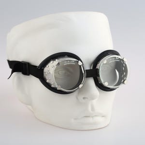 May include: Steampunk-style goggles featuring clear circular lenses, silver-toned metal side shields with a studded texture, and a black padded frame. The goggles are secured with an adjustable black elastic strap and are displayed on a white mannequin head.