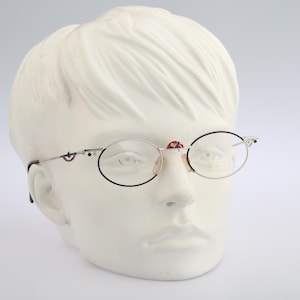 May include: Silver-framed eyeglasses with round lenses and black rims are displayed on a white mannequin head. The glasses have decorative red and black accents on the bridge and temples. The overall design is vintage and artistic.