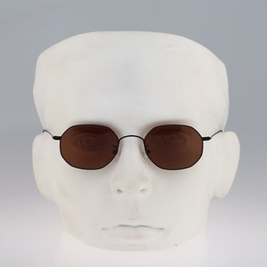May include: A pair of black octagon-shaped sunglasses with brown lenses. The sunglasses are on a white mannequin head.