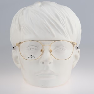 May include: Gold-tone metal eyeglasses with a double bridge design and thin frames. The clear lenses feature the Gucci brand name and a small logo printed in black on the lower left side. The glasses are displayed on a white mannequin head.