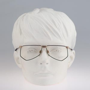 May include: Spectacles featuring a thin, black metal frame with a distinct geometric, angular shape. The bridge and temple attachments are finished in a metallic gold tone. The clear lenses are set within the dark rims, creating a sharp contrast.