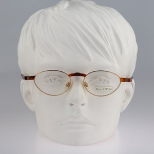 Op de afbeelding: Een paar ovale brillen met een bruin schildpad montuur en gouden metalen accenten. De bril zit op een wit mannequins hoofd. De merknaam "Marc O'Polo" is zichtbaar op de rechter lens.