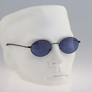 Puede incluir: Gafas de sol ovaladas con montura fina de metal negro y cristales tintados en azul oscuro. Las gafas se muestran sobre una cabeza de maniquí blanca, destacando las varillas delgadas y el puente que une los dos aros circulares de las lentes.
