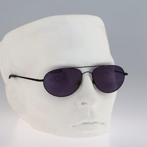 May include: Aviator sunglasses featuring a thin black metal frame and dark tinted lenses. The eyewear has a classic double bridge design with slender temples, providing a sleek and minimalist look for everyday use with a neutral aesthetic.