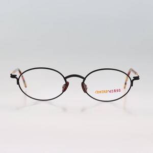 May include: Black oval-shaped eyeglasses with a thin metal frame. The brand name "CONTROVERSE" is printed in red and orange on the right lens.