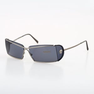 Könnte beinhalten: Sonnenbrille mit silbernem Rahmen und dunkelgrauen Gläsern. Die Sonnenbrille hat eine einzigartige, eckige Form und ein kleines Logo an der Seite.