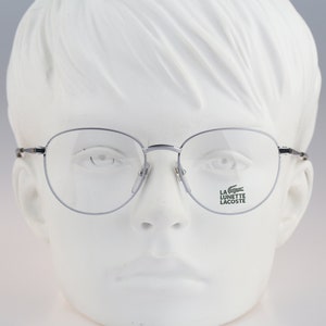 Puede incluir: Gafas de montura plateada con lentes transparentes. El logotipo de La Lunette Lacoste está impreso en la lente derecha.