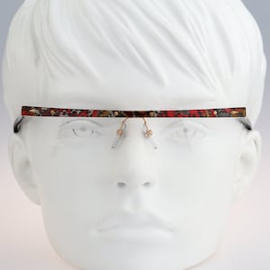 Puede incluir: Gafas sin montura con una barra superior recta y plana con un diseño detallado en rojo, negro y dorado. El marco incluye almohadillas nasales y componentes de puente de metal en tono dorado, mostrando una estética horizontal y elegante.
