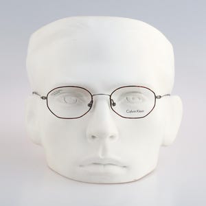 May include: Calvin Klein eyeglasses featuring a thin, dark metal frame with a rounded hexagonal shape. The lenses are clear, and the brand name Calvin Klein is printed in black on the right lens. The glasses are displayed on a white mannequin head.