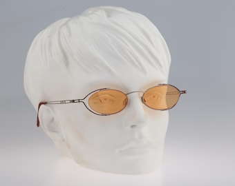 May include: Oval-shaped glasses with thin silver-tone metal frames and light orange-tinted lenses. The frames feature a distinctive double-rim design around the lenses and slender temples with a small rectangular detail near the hinges.