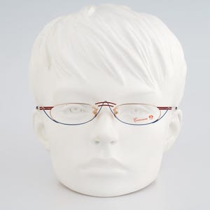 May include: Gold-toned eyeglasses with red and blue accents are displayed on a white mannequin head. The glasses have a delicate frame and oval lenses. The brand name is printed on the right lens.