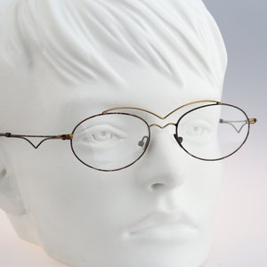 May include: A pair of oval-shaped eyeglasses with a thin gold metal frame and clear lenses. The glasses are on a white mannequin head.